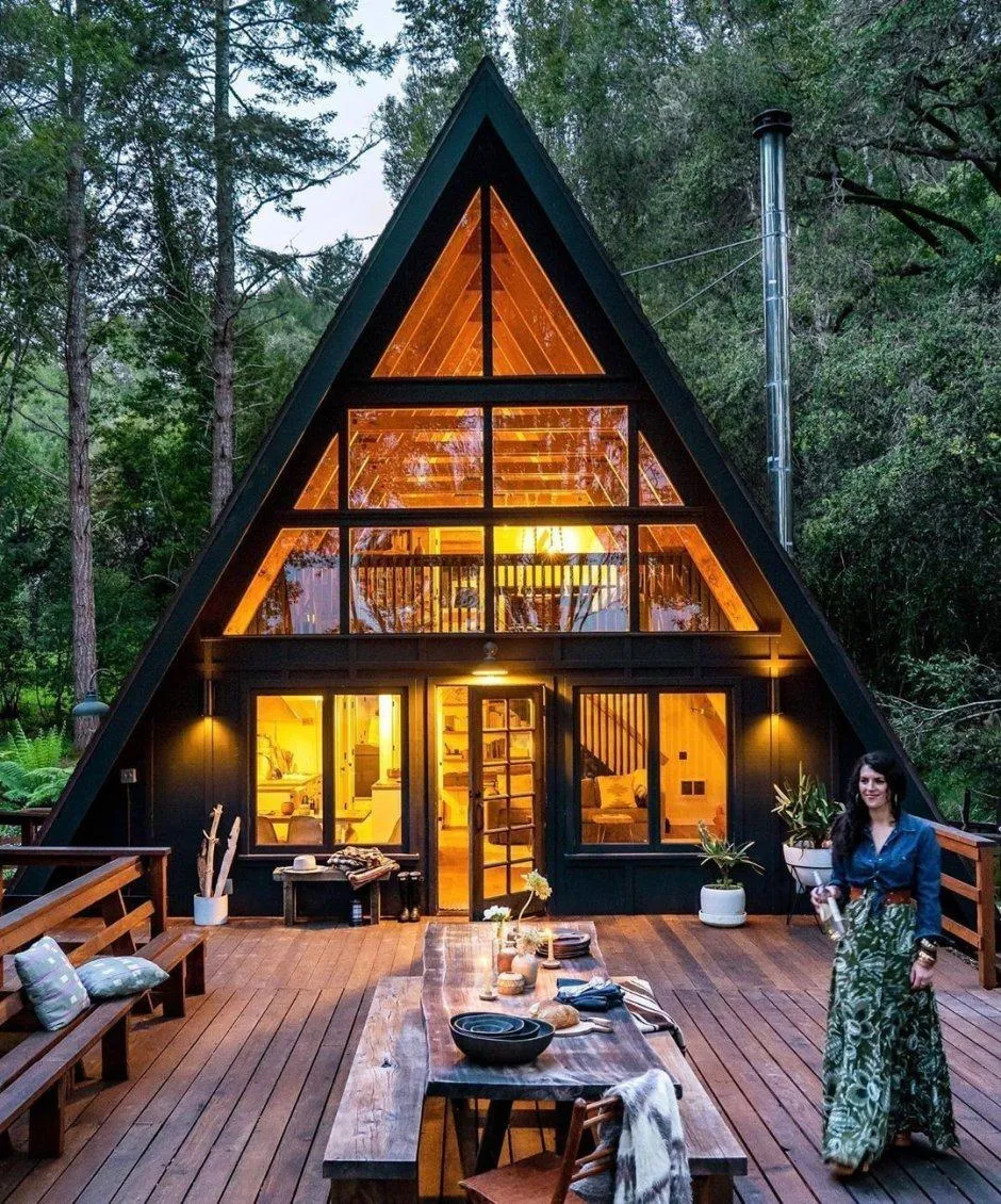 Дом A-Frame в лесу вечером: светящееся треугольное остекление и терраса